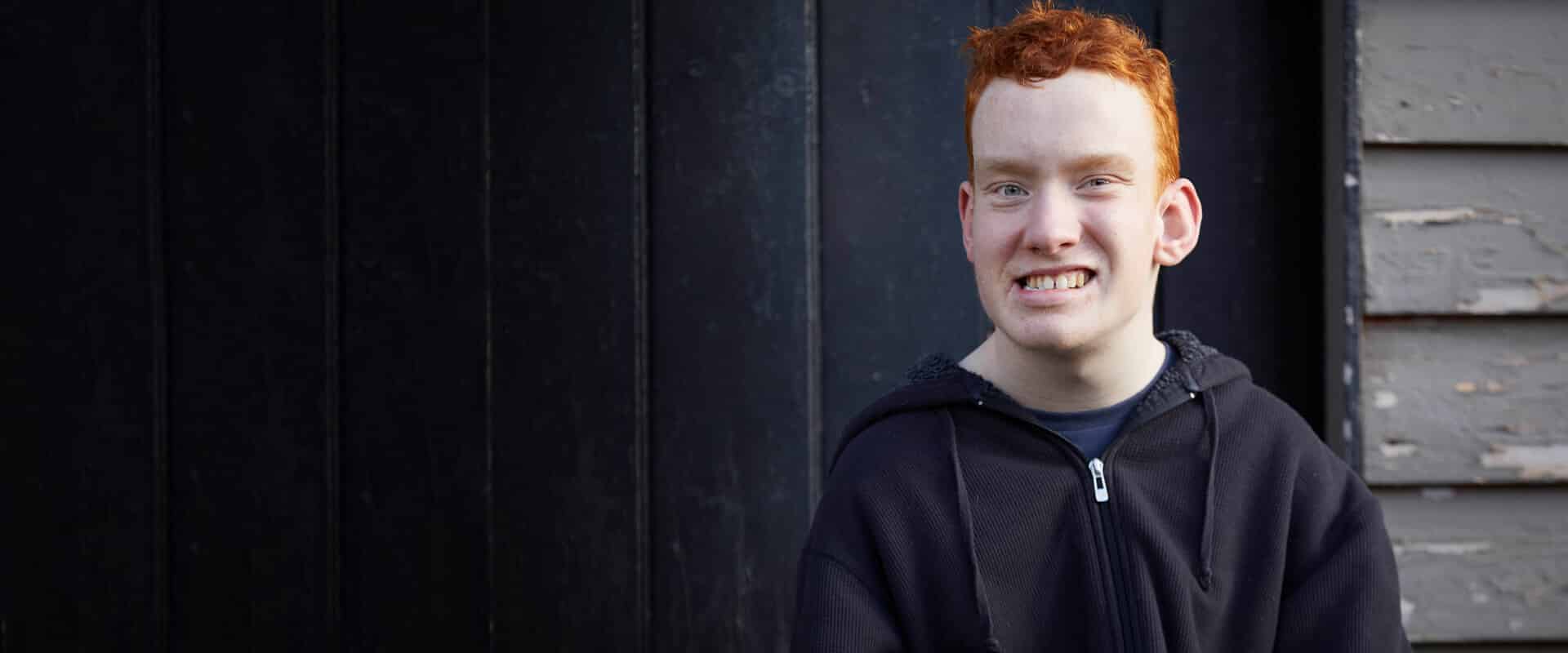 Smiling young man with red hair wearing a black hoodie, standing in front of a dark wooden wall and weathered grey siding.
