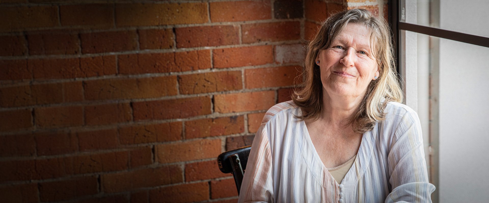 Middle-aged woman with a gentle smile sits by a window in front of a brick wall, wearing a light blouse and looking relaxed.